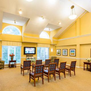 A brightly lit living room with a row of chairs facing a television, framed by large windows and yellow walls.