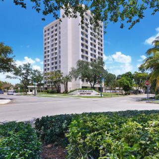 A tall, white high-rise building surrounded by lush greenery and trees under a bright blue sky.