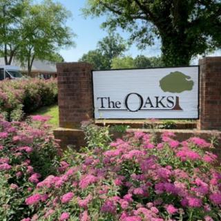 A scenic view of a sign for 'The Oaks' surrounded by vibrant flowers and trees.