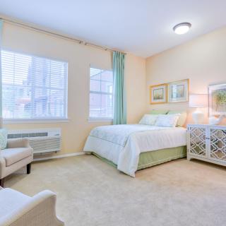 A cozy and elegantly decorated bedroom with light-colored walls, featuring a comfortable bed, chair, and windows with natural light.
