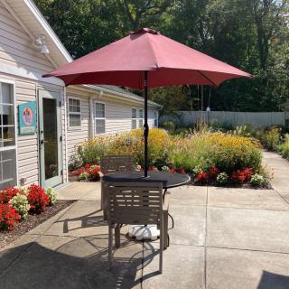 A sunny patio with a table and chairs under a red umbrella, surrounded by vibrant flowers.