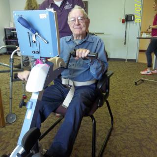 An elderly person using a stationary exercise machine in a healthcare setting.