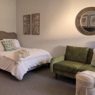 A cozy bedroom with a single bed and a green armchair, accented by a round mirror and decorative wall art.