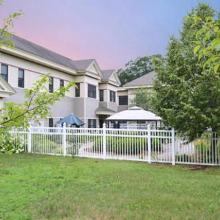 A view of a two-story residential building with a well-maintained lawn and white fence, featuring outdoor furniture under umbrellas.