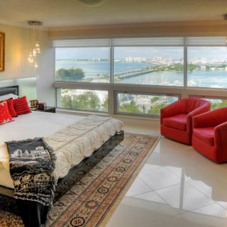 A modern bedroom with large windows overlooking a scenic ocean view, featuring elegant decor and red accent chairs.