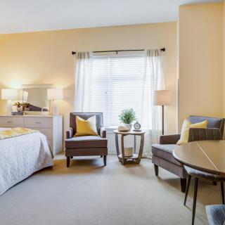 A cozy bedroom featuring a comfortable bed, two armchairs, and a small table near a window with sheer curtains.