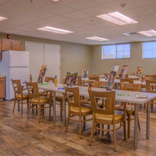 A well-lit dining room with several wooden tables and chairs, featuring a refrigerator in the corner.
