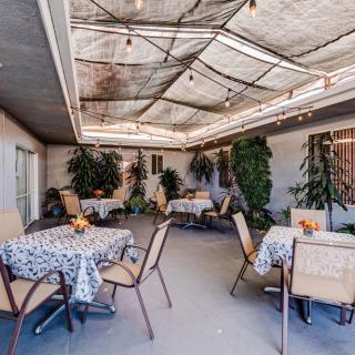A cozy outdoor patio with tables, chairs, and string lights under a covered area surrounded by greenery.