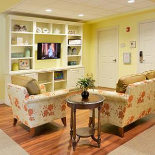 A cozy living room with floral armchairs facing a wall-mounted television, surrounded by decorative shelves and a central table with a plant.
