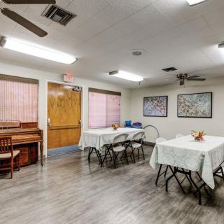 A community room with tables and chairs set up, featuring a piano against the wall and fluorescent lighting.