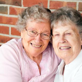 Two elderly women smiling and hugging in front of a brick wall.