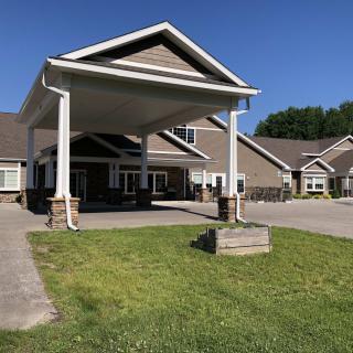 A building entrance with a canopy, surrounded by a driveway and grassy lawn on a sunny day.