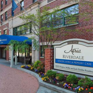 Entrance of a building with a blue awning and prominent signage, surrounded by flowers and trees.
