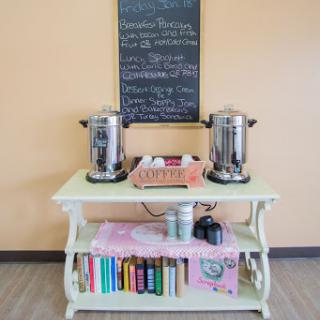 A cozy coffee station featuring beverage dispensers and a chalkboard menu, complemented by colorful books on a decorative shelf.