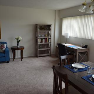 A cozy living and dining space featuring a bookshelf, desk with a lamp, dining table set for two, and comfortable seating.
