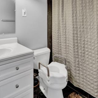 A bathroom featuring a toilet with a handrail for accessibility, a sink, and a patterned shower curtain.