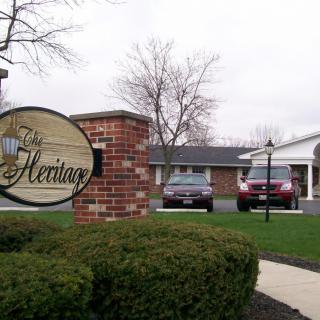 A view of a brick sign labeled 'The Heritage' in front of a building with two cars parked nearby.