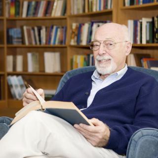 A man with glasses is sitting in a library, relaxed and reading a book.