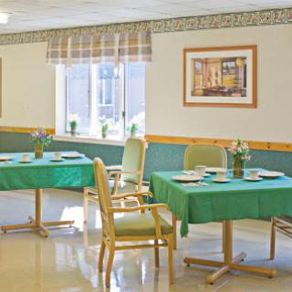 A bright and inviting dining area featuring tables with green tablecloths, comfortable chairs, and floral arrangements near the windows.