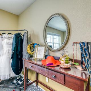 A dressing room featuring a mirror, clothing rack with a wedding dress and suit, and a table adorned with hats, ties, and decorative items.