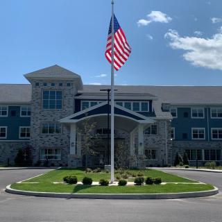 A large building with an American flag prominently displayed in front, surrounded by well-maintained landscaping and a clear blue sky.