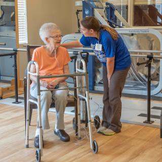 A caregiver assists an elderly woman using a walker in a rehabilitation setting.