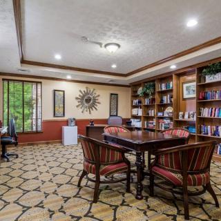A cozy library room with bookshelves, a central table with chairs, and large windows providing natural light.