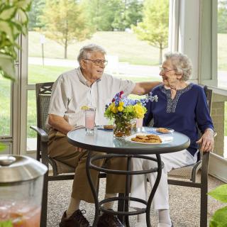 An elderly couple enjoys a pleasant conversation at an outdoor table surrounded by nature.