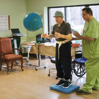 A person undergoing physical therapy with the assistance of a healthcare professional, practicing balance on a foam pad.