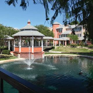 A serene outdoor scene featuring a gazebo by a pond with a fountain, set against the backdrop of a charming building.