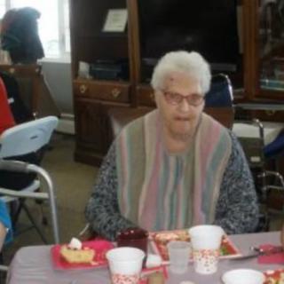 An elderly woman with glasses enjoying a meal indoors, seated at a table set for a celebration.
