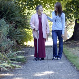 An elderly woman walking with a caregiver in a park setting.