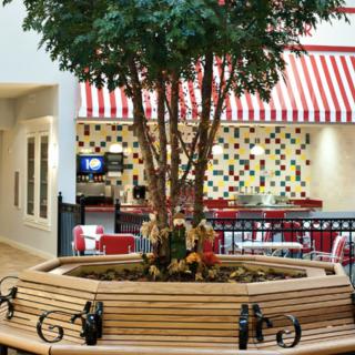 A cozy indoor seating area with benches surrounding a central tree in a mall's food court, featuring striped awnings and colorful tiles.