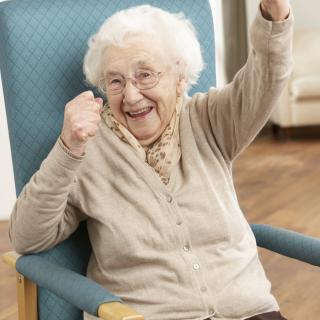 A joyful elderly woman celebrating while seated in a chair indoors.