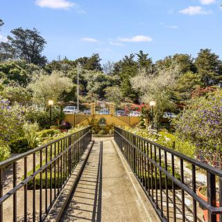 A scenic pathway bridge leading through a lush garden filled with trees and greenery under a clear blue sky.
