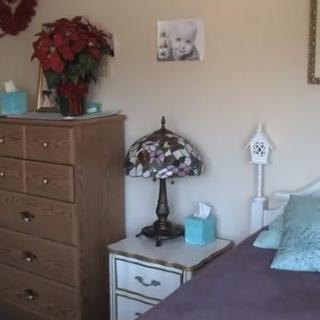 A cozy bedroom featuring a wooden dresser, nightstand with a decorative lamp, and floral decorations.
