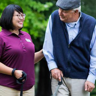 An elderly man walking outdoors with the support of a caregiver.