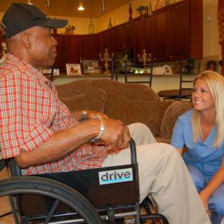 A caregiver kneels next to a man in a wheelchair, engaging in conversation.