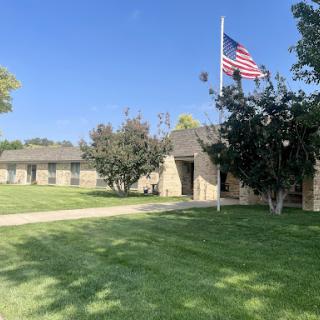 A peaceful view of a building with an American flag, surrounded by well-kept grass and trees under a clear blue sky.