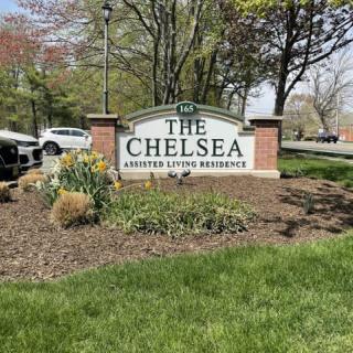 Sign for The Chelsea Assisted Living Residence surrounded by landscaped greenery.