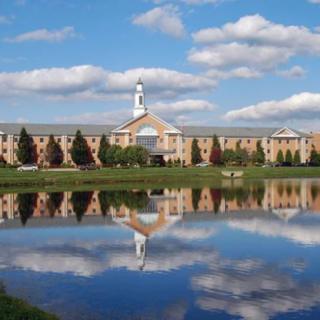 A large building reflected in a calm lake under a partly cloudy sky.