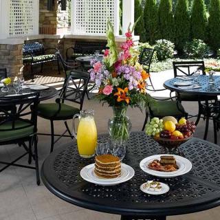 A cozy outdoor breakfast setting with pancakes, orange juice, and fresh fruit on a patio table adorned with colorful flowers.