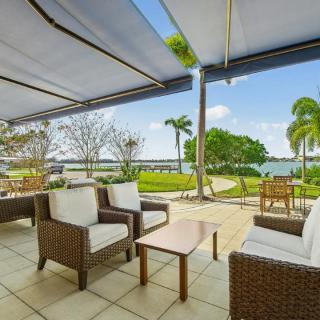 A serene patio with comfortable wicker seating overlooks a tranquil lake surrounded by lush greenery and palm trees.