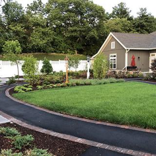 A well-maintained garden with a lush lawn, trees, and a neat driveway in front of a house.