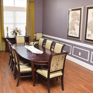 A well-decorated dining room featuring a long wooden table with upholstered chairs, framed artwork on lavender walls, and hardwood flooring.