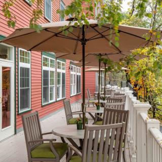 A cozy outdoor patio with tables and umbrellas on a terrace beside a red building, surrounded by lush greenery.