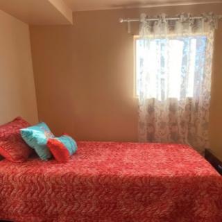 A cozy bedroom with a red bedspread and colorful pillows next to a window with sheer curtains.
