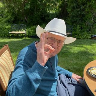 An elderly man wearing a cowboy hat and glasses makes an 'OK' gesture while sitting outdoors.