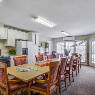 A spacious dining area with a long table, surrounded by chairs and adjoining a well-lit kitchen.