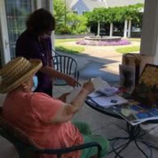 An elderly person wearing a hat interacts with a caregiver on an outdoor patio.